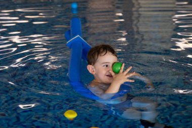 Mutlu çocuk mavi yüzme cihazıyla havuzda yüzmeyi öğrenirken renkli toplarla oynarken eğleniyor.