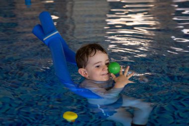 Küçük çocuk yüzme derslerini seviyor, kapalı havuzda şişme kolluk takarken renkli toplarla oynuyor.