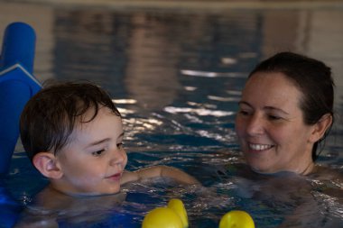 Küçük çocuk yüzme hocasıyla yüzmeyi ve yüzme cihazını kapalı havuzda öğreniyor, eğleniyor ve suya güven inşa ediyor.