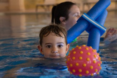 Genç çocuk, su güvenliği ve erken çocukluk gelişimi konularında eğitmen ve renkli oyuncaklarla yüzme dersinin keyfini çıkarıyor.