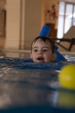 Küçük çocuk yüzme derslerini seviyor, sarı bir topla oynarken kapalı bir havuzda mavi bir yüzme aleti takıyor.
