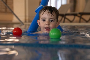 Genç çocuk yüzme dersini seviyor, renkli toplarla oynarken kapalı havuzda yüzme aleti kullanıyor.