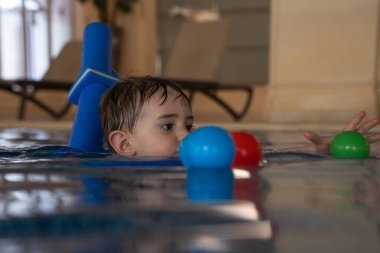 Küçük çocuk yüzme dersleri almaktan, yüzme aleti kullanmaktan ve kapalı havuzda renkli toplarla oynamaktan hoşlanıyor.