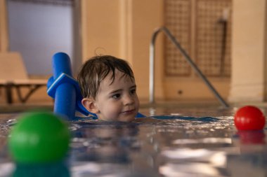 Küçük çocuk yüzme derslerini seviyor, yüzme aygıtı ve kapalı havuzda renkli toplar kullanıyor.