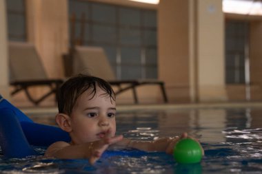 Küçük çocuk yüzme dersleri alıyor, otel havuzunda renkli balolara uzanıyor, su güvenliğini ve erken çocukluk gelişimini destekliyor