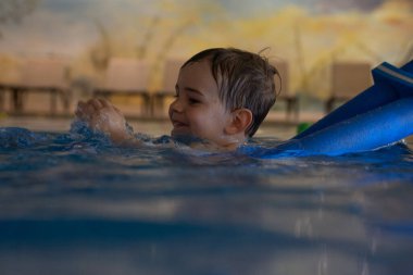 Kapalı havuzda mavi bir yüzme cihazıyla yüzmeyi öğrenen, su sıçratan ve oynayan mutlu çocuk.