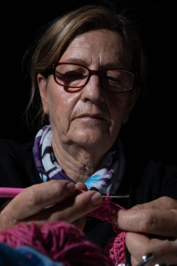 Odaklanmış son sınıf öğrencisi bir kadın kancayla pembe iplik örmeye odaklanıyor, loş ışıklandırılmış bir ayarda dokulu bir parça yaratıyor.