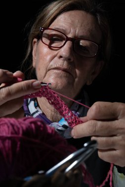 Pembe iplikle ustalıkla örgü örüp loş bir ortamda karmaşık dikişler atan son sınıf öğrencisi bir kadın.