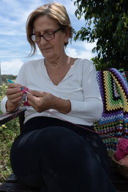 Bahçesinde dinlenen yaşlı bir kadın, huzurlu bir yaz öğleden sonra eğlenirken canlı bir el işi tığ işi yapıyor.