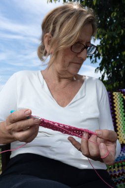 Yaşlı bir kadın rahatlatıcı bir hobi ediniyor, açık havada pembe iplikler örüyor, geleneksel el sanatları ve aktif emeklilik sanatları sergiliyor.