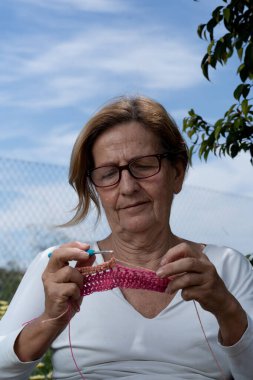 Yaşlı bir kadın açık havada pembe bir kumaş parçası örmeye odaklanıyor, doğada barışçıl bir hobi ediniyor.