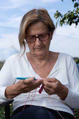 Yaşlı bir kadın arka bahçesinde huzurlu bir öğleden sonranın tadını çıkarıyor. Yumuşak pembe ipliklerle örgü örüyor, en sevdiği boş zamanlarını kucaklıyor.
