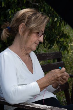 Yaşlı bir kadın, yemyeşil bir yemlikle çevrili ahşap bir bankta renkli iplikler örerek rahatlatıcı bir öğleden sonranın tadını çıkarıyor.