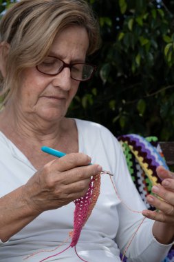 Kancayla renkli iplikler örmeye odaklanan yaşlı bir kadın, dışarıda huzurlu bir ortamda el yapımı el sanatları yapıyor.
