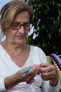 Kancayla renkli iplikler örmeye odaklanan yaşlı bir kadın, dinlendirici bir açık hava aktivitesinin keyfini çıkarıyor.