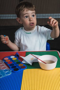 Suluboya boyayla resim yapan ve renkli masada otururken parmağını kaldıran genç bir çocuk.