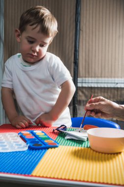 Meraklı çocuk renkli bir masada canlı suluboya resim yapan öğretmeni dikkatle izliyor, eğlenceli bir öğrenme deneyimi yaşıyor.