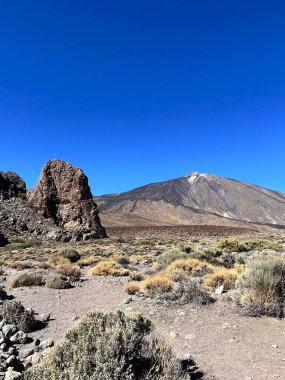 Bereketli mavi gökyüzünün altında yükselen Teide Dağı ile volkanik arazinin manzarası.