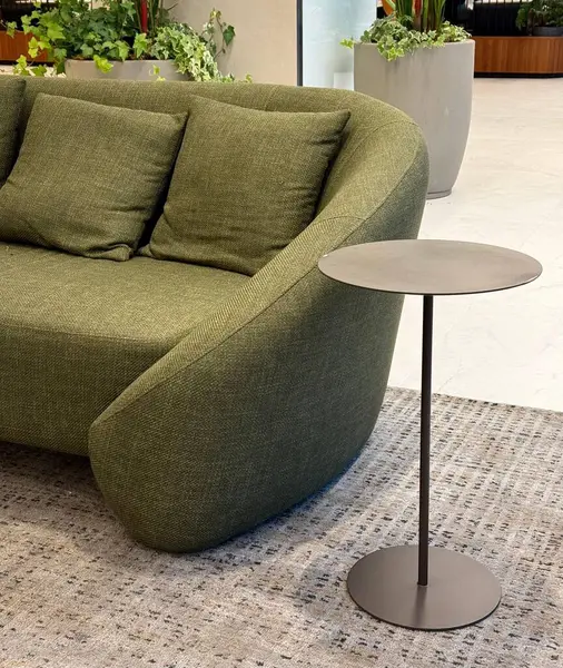 Modern, iyi ışıklandırılmış yeşil bitkilerle çevrili, parlak minimalist bir metal yan sehpanın yanında, yüksek sırtı ve desenli döşemeleriyle çağdaş zeytin rengi bir kanepe..