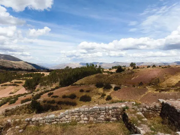 Yuvarlanan tepeler ve ekilmiş topraklar And bölgesindeki parlak, parçalı bulutlu bir gökyüzünün altında uzanıyor. Antik taş yapılar bu arazide zengin bir tarihi işaret ediyor..