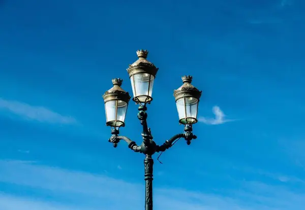 Bu resimde güzel dizayn edilmiş üç lamba direği ince bulutlu canlı mavi gökyüzüne karşı duruyor. Lambaların süslü detayları biraz klasik zarafet katıyor..