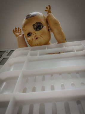 Zarar görmüş eski model bir oyuncak bebek ürkütücü bir şekilde beyaz bir plastik tepsinin üzerine bakar..
