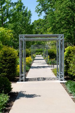 Geometrik metal kemerler, yeşillik ve düzgünce budanmış çalılarla çevrili, peyzajlı bir bahçede temiz bir beton yolu kaplıyor. Güneşli bir günde doğal ışık ve derin bir perspektifle yakalandı..