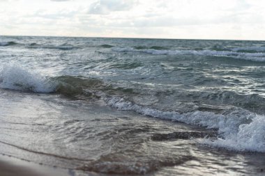 Islak kumsala düşen göl dalgalarının yakın görüntüsü. Fırtınalı göl suyu, dalgaların ve köpüklerin detaylı dokusuyla ıslak kumun üzerinde süzülüyor. Michigan Gölü üzerinde bulutlu gökyüzünün altında doğal manzaralı kıyı şeridi ele geçirildi.