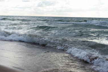 Islak kumsala düşen göl dalgalarının yakın görüntüsü. Fırtınalı göl suyu, dalgaların ve köpüklerin detaylı dokusuyla ıslak kumun üzerinde süzülüyor. Michigan Gölü üzerinde bulutlu gökyüzünün altında doğal manzaralı kıyı şeridi ele geçirildi.