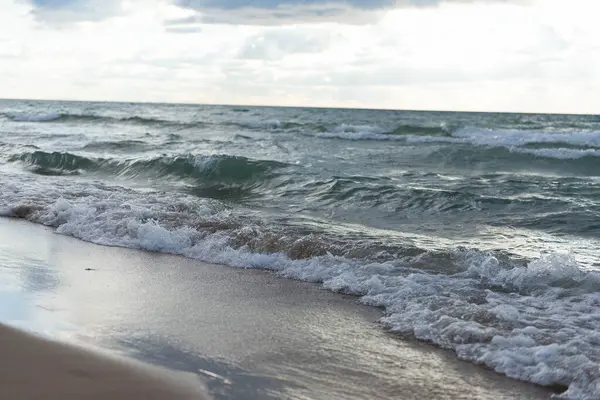 Islak kumsala düşen göl dalgalarının yakın görüntüsü. Fırtınalı göl suyu, dalgaların ve köpüklerin detaylı dokusuyla ıslak kumun üzerinde süzülüyor. Michigan Gölü üzerinde bulutlu gökyüzünün altında doğal manzaralı kıyı şeridi ele geçirildi.