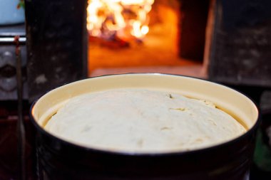 Yanan odun odunuyla geleneksel ahşap fırının yanına yerleştirilmiş diş minesi kabına yükselen maya hamuru yakın plan. Kırsal bir mutfakta kırsal ekmek yapma süreci ev yapımı pişirmenin sıcaklığını ve sıcaklığını yakalıyor. Ukrayna aşçılık geleneği