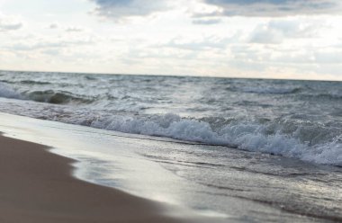 Gün batımında bulutlu gökyüzünün altında kumlu kıyıları yıkayan köpüklü dalgalarla huzurlu deniz manzarası. Sakin deniz suyuyla doğal kıyı manzarası.