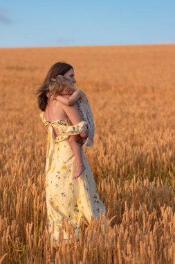 Gün batımında küçük kızını tarlada tutan güzel bir bayan. Güzel sarı elbiseli çocuklu bir kadın gün batımında tarlada poz verir..