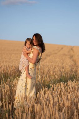 Gün batımında küçük kızını tarlada tutan güzel bir bayan. Güzel sarı elbiseli çocuklu bir kadın gün batımında tarlada poz verir..