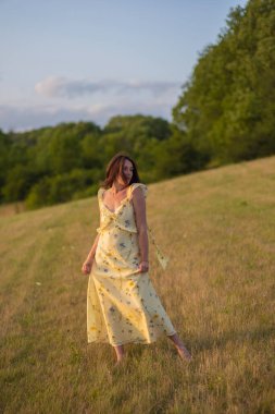 Genç ve güzel bir bayan gün batımında çıplak ayakla bir tarlada yürüyor. Güzel sarı elbiseli bir kız gün batımında tarlada poz verir..
