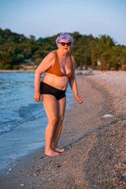 Güneş gözlüklü ve mayolu yaşlı bir kadın sahilde deniz kıyısında yürüyor. Avrupa görünümlü, mor saçlı bir kadın denizin, ormanın ve dağların arka planında.