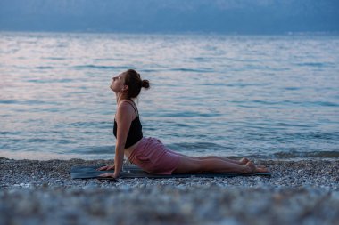 Siyah kolsuz tişörtlü ve pembe şortlu genç bir kız deniz sahilinde yoga yapıyor. Güzel Adriyatik Denizi 'ne ve yeşil kıyılara bakan gün batımında yoga. Fotoğraf sağlıklı bir yaşam tarzını, sporu, yogayı teşvik eder.