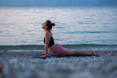 Siyah kolsuz tişörtlü ve pembe şortlu genç bir kız deniz sahilinde yoga yapıyor. Güzel Adriyatik Denizi 'ne ve yeşil kıyılara bakan gün batımında yoga. Fotoğraf sağlıklı bir yaşam tarzını, sporu, yogayı teşvik eder.