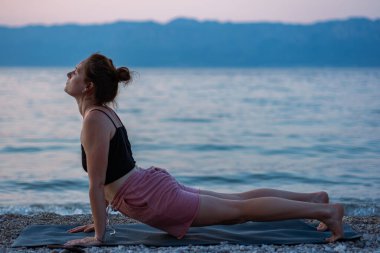 Siyah kolsuz tişörtlü ve pembe şortlu genç bir kız deniz sahilinde yoga yapıyor. Güzel Adriyatik Denizi 'ne ve yeşil kıyılara bakan gün batımında yoga. Fotoğraf sağlıklı bir yaşam tarzını, sporu, yogayı teşvik eder.