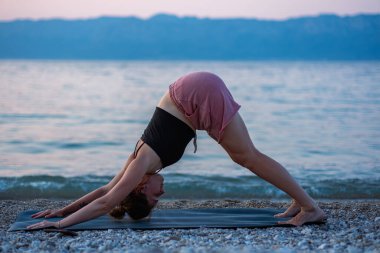 Siyah kolsuz tişörtlü ve pembe şortlu genç bir kız deniz sahilinde yoga yapıyor. Güzel Adriyatik Denizi 'ne ve yeşil kıyılara bakan gün batımında yoga. Fotoğraf sağlıklı bir yaşam tarzını, sporu, yogayı teşvik eder.
