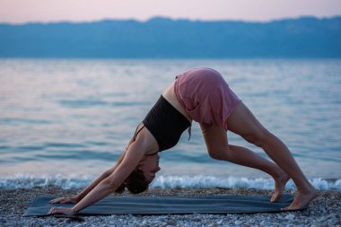 Siyah kolsuz tişörtlü ve pembe şortlu genç bir kız deniz sahilinde yoga yapıyor. Güzel Adriyatik Denizi 'ne ve yeşil kıyılara bakan gün batımında yoga. Fotoğraf sağlıklı bir yaşam tarzını, sporu, yogayı teşvik eder.