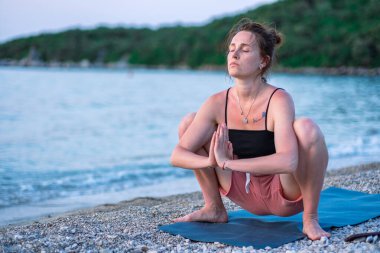 Siyah kolsuz tişörtlü ve pembe şortlu genç bir kız deniz sahilinde yoga yapıyor. Güzel Adriyatik Denizi 'ne ve yeşil kıyılara bakan gün batımında yoga. Fotoğraf sağlıklı bir yaşam tarzını, sporu, yogayı teşvik eder.