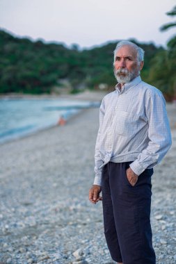 Plajda beyaz gömlekli ve siyah pantolonlu yaşlı bir adam. Sakallı yaşlı bir adam gün batımında sahilde yalnız yürür. Yalnız, gri saçlı yaşlı bir adamın portresi. Arka planda palmiye ağaçları.
