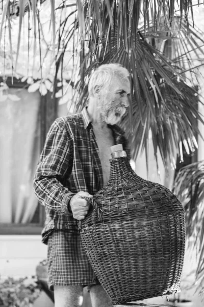 一位老酿酒师摆设着一个用藤条包裹在棕榈树下的老式大酒瓶 俊俏迷人的祖父手里拿着一只装着葡萄的瓶子 — 图库照片