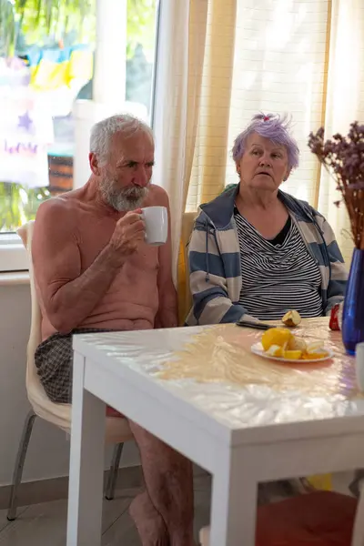 An elderly woman and an old man with a beard are sitting at a table in the living room and drinking tea. An elderly couple is resting in their bright house.