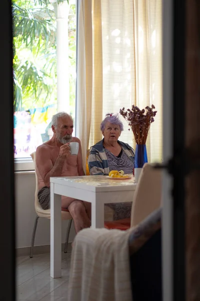 An elderly woman and an old man with a beard are sitting at a table in the living room and drinking tea. An elderly couple is resting in their bright house.