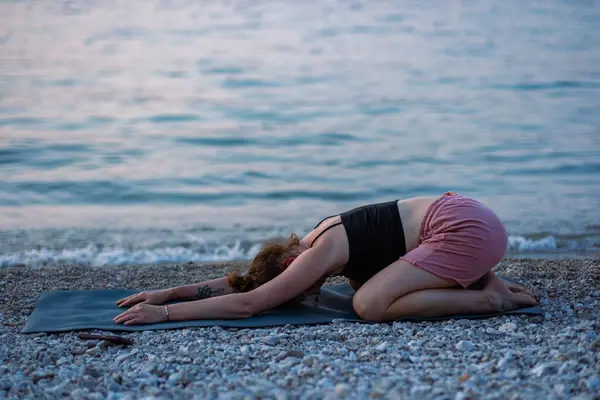 Siyah kolsuz tişörtlü ve pembe şortlu genç bir kız deniz sahilinde yoga yapıyor. Güzel Adriyatik Denizi 'ne ve yeşil kıyılara bakan gün batımında yoga. Fotoğraf sağlıklı bir yaşam tarzını, sporu, yogayı teşvik eder.