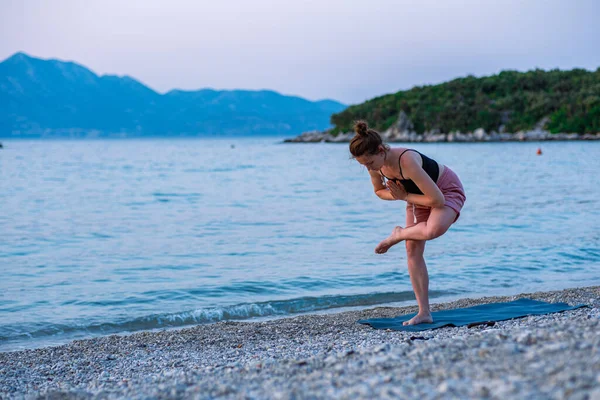 stock image A young girl in a black tank top and pink shorts doing yoga on the sea beach. Yoga at sunset overlooking the beautiful Adriatic Sea and green shores. Photo promotes a healthy lifestyle, sports, yoga.