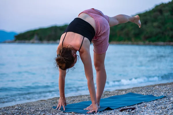 stock image A young girl in a black tank top and pink shorts doing yoga on the sea beach. Yoga at sunset overlooking the beautiful Adriatic Sea and green shores. Photo promotes a healthy lifestyle, sports, yoga.