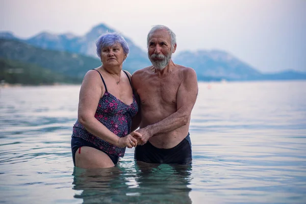 Elderly couple swims in the sea. Old bearded man and aged woman in swimsuits together stand in the tranqise water of Adriatic sea at sunset.